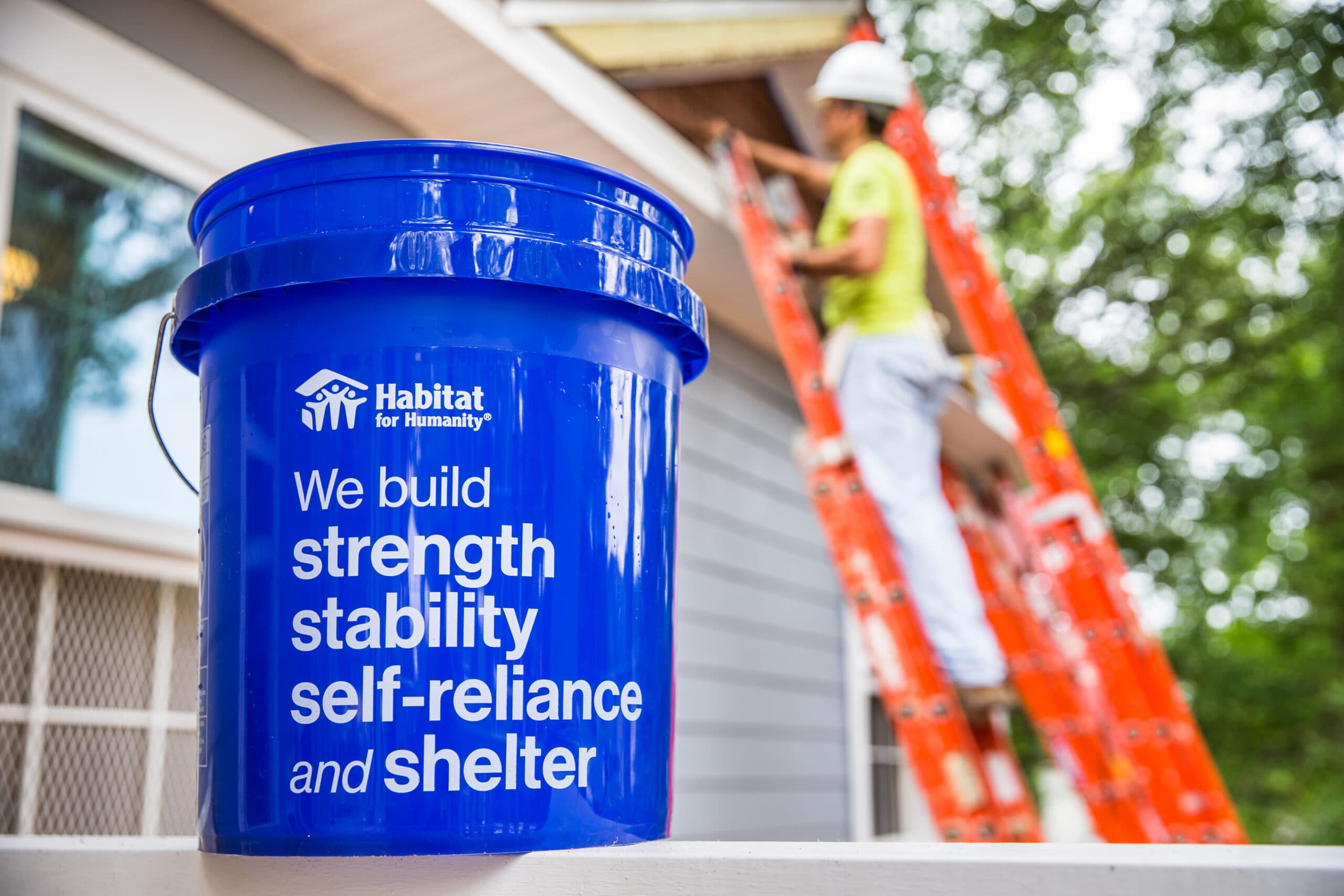 A Habitat for Humanity bucket reading 'We build strength, stability, self-reliance and shelter' at a build site