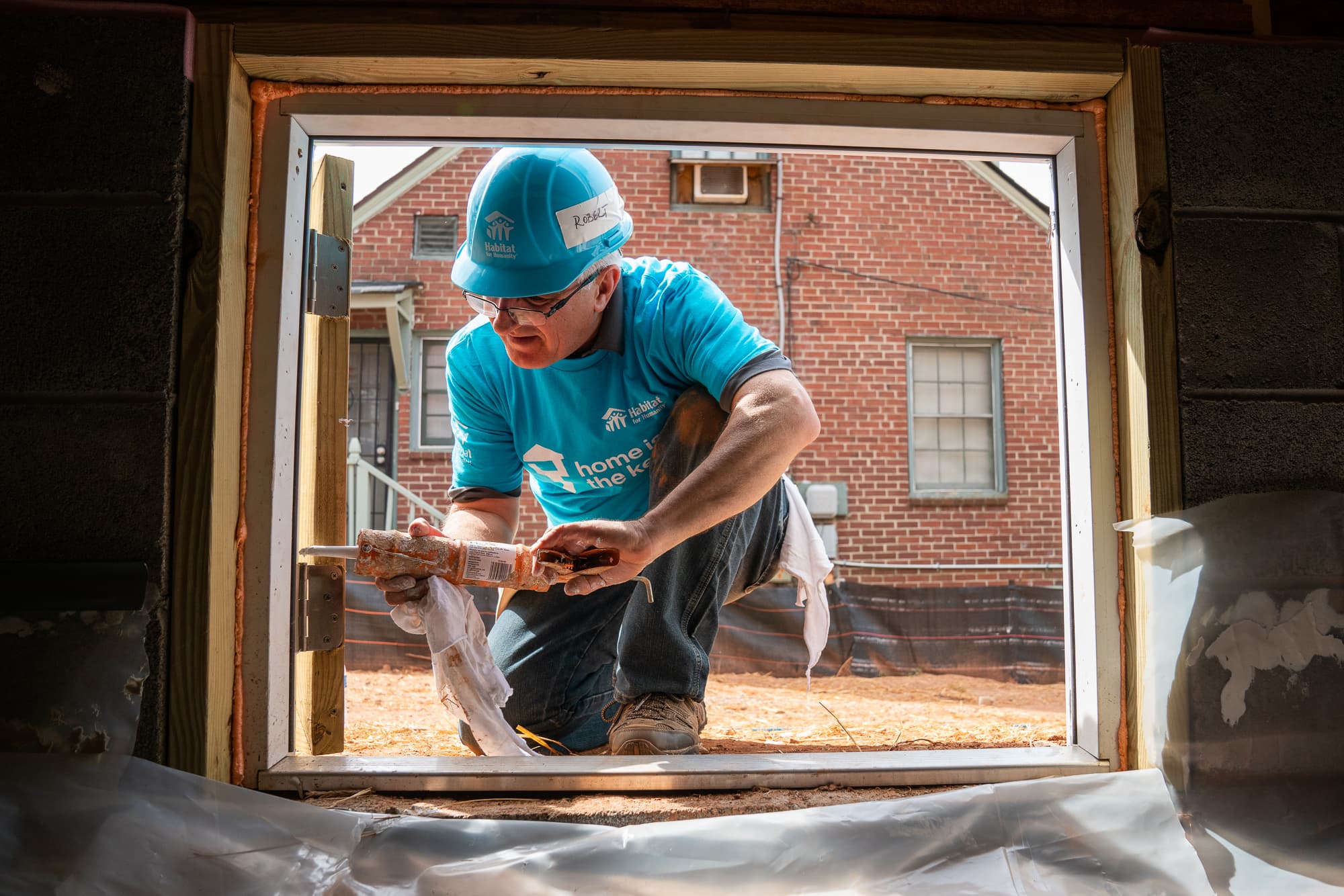 A volunteer in a 'Home is the Key' shirt framing a window opening on a Habitat build